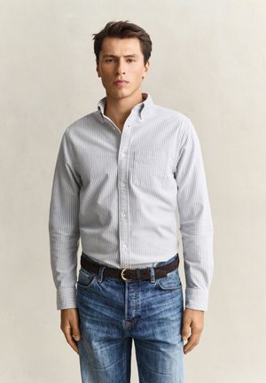 Jeune homme aux cheveux bruns courts portant une chemise légère rayée boutonnée rentrée dans un jean bleu avec une ceinture marron foncé, debout devant un fond uni.