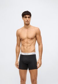 Mužský model se svalnatou postavou nosí černé vzorované boxerky Calvin Klein na čistém světlém pozadí.