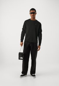 Svart sweatshirt med rund hals, tillsammans med vida svarta jeans. Håller en liten svart The North Face-väska. Svarta solglasögon fulländar stilen.