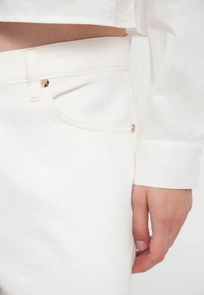 Un pantalon en denim blanc présente des accessoires en ton doré, une coupe slim et une taille légèrement montée, associé à une chemise blanche à boutons.