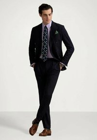 Navy pinstripe suit with double-breasted jacket, light purple shirt, patterned tie, and brown leather shoes. Green pocket square detail.