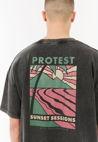 Zwarte oversized t-shirt met een grafisch ontwerp in pastelkleuren. De tekst luidt "PROTEST" en "SUNSET SESSIONS" met een strandscène en palmboom.