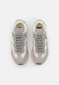 Ténis atléticos cinza e creme, feitos de malha e camurça, com atacadores planos, bico redondo e sola de borracha. Apresenta um logótipo azul.