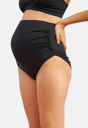 Žena nosiace čierne bikiny nohavičky s vysokým pásom a bočnými záhybmi, pózujúca s jednou rukou opretou o bok.