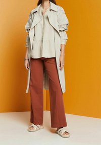 Trench beige sobre una blusa peplum de color crema claro; combinada con pantalones de pierna ancha en color óxido y sandalias blancas de tiras. Fondo de color sólido.