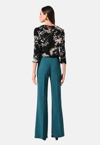 Zwarte top met bloemenprint, driekwartmouwen en een keyhole-detail aan de achterkant, gecombineerd met tealgroene wijde broek. Platte schoenen zichtbaar.