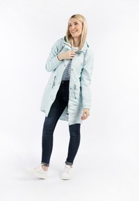Giacca impermeabile blu chiaro con cappuccio, caratterizzata da lati arricciati e ricami decorativi, abbinata a jeans scuri e sneakers bianche.