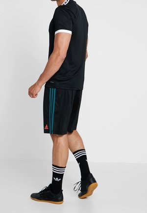 Mężczyzna w czarnym stroju sportowym z turkusowymi paskami na spodenkach, rękawami wykończonymi białym kolorem, czarnych skarpetach Adidas i czarnych halowych butach piłkarskich.