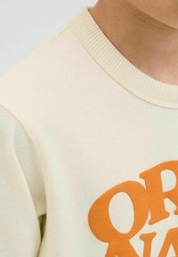 Gai bēšs sporta krekls ar ribinātu apkakli. Priekšpusē ir izteikts oranžs grafiskais teksts. Materiāls ir rāmā tekstūra.