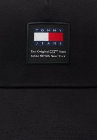 Gorra negra con un diseño estructurado. Presenta un parche cuadrado con el texto "TOMMY JEANS" y un logo en rojo, blanco y azul. El material parece suave.