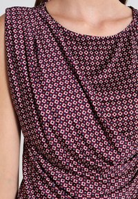 Czerwono-granatowy wzorzysty top z miękkim drapowaniem, z geometrycznymi kształtami diamentów i plisowanym detalem na ramieniu. Gładka tekstura.