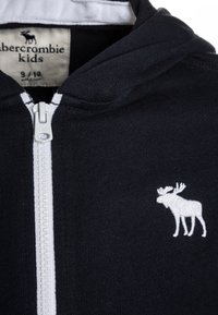 Zwarte zip-up hoodie van zachte stof, met een witte rits en een geborduurd moose-logo op de borst. Maat 9/10.