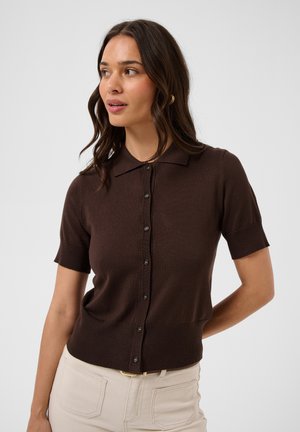 Femme aux longs cheveux foncés portant un haut brun à manches courtes et boutonné, et un pantalon beige clair, regardant vers la gauche sur un fond uni.