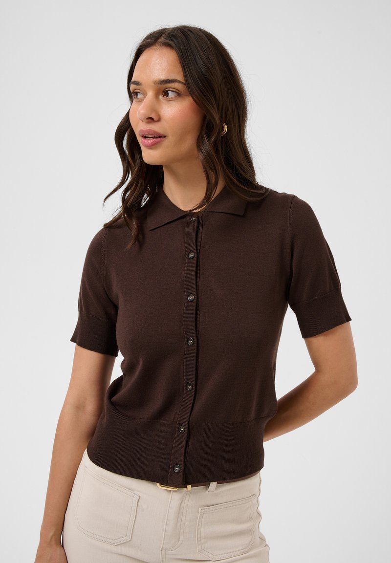 Femme aux longs cheveux foncés portant un haut brun à manches courtes et boutonné, et un pantalon beige clair, regardant vers la gauche sur un fond uni.