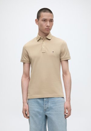 Man wearing beige short-sleeve polo shirt with small logo on chest and light blue jeans, standing against a plain white background.