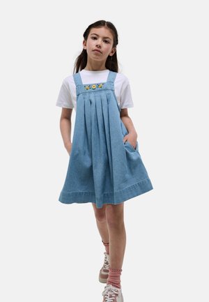 Fille portant une robe en jean bleue plissée avec broderie florale sur un t-shirt blanc, des chaussettes roses et des baskets blanches, debout les mains dans les poches.