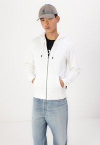 Witte rits-hoodie met voorzakken, trekkoordcapuchon en zwarte ritshardware. Ggedragen over een zwart T-shirt en lichtblauwe jeans.