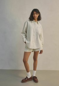Přesně padnoucí, světle béžová pruhovaná košile s límcem a přední kapsou, doplněná o krátké šortky stejné barvy. A outfit doplňují hnědé boty a bílé ponožky.