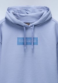 Lyseblå hettegenser med en frontlomme, ribbestrikkede mansjetter, snor og en blå grafisk logo med påskriften "NAPAPIRI geographic." Myk tekstur.