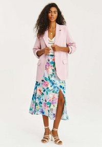 Světle růžový blejzr, bílý saténový top, maxi sukně s květinovým vzorem a bočním rozparkem, a bílé lodičky s kotníkovým páskem. Texturované tkaniny a živé barvy.