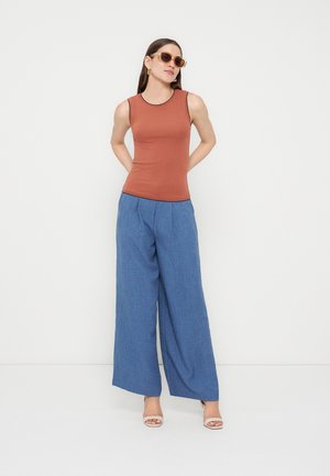 Γυναίκα με γυαλιά ηλίου, αμάνικη μπλούζα χρώματος σκουριάς, ευρύχωρο μπλε παντελόνι και μπεζ πέδιλα με ανοιχτά δάχτυλα, στέκεται μπροστά σε λευκό φόντο.