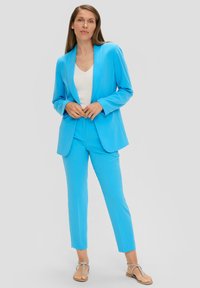 Blazer ajusté bleu clair et pantalon assorti en tissu léger. Design simple avec des poches, associé à un haut blanc et des sandales.