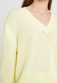 Jasnożółty sweter z dzianiny z dekoltem w serek, luźnym krojem oraz pionowym paskowanym wzorem i ażurowym splotem.