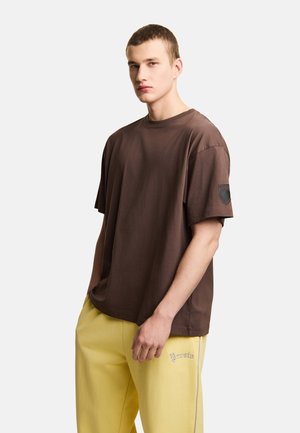 Młody mężczyzna stoi z rękami w kieszeniach, ubrany w oversize brązowy T-shirt i żółte spodnie z logo na lewym udzie.