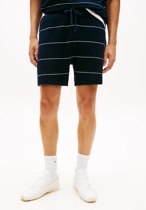 Muž oblečený v námornícky modrých pletených šortkách s bielymi horizontálnymi pruhmi, bielych členkových ponožkách a bielych teniskách, stojaci pred bielym pozadím.