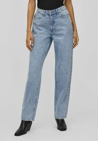 Heledad sinised kõrge vöökohtadega teksad sirge säärega, tekstuuriga denim, viie taskuga disain ja nööpidega sulgur, koos mustade kontsakingadega.