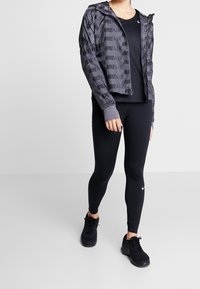 Harmaa kuvioitu vetoketjullinen takki, jossa on "NIKE AIR" -logo, yhdistettynä mustiin leggingseihin ja mustiin urheilujalkineisiin. Sujuva kangas, joka on istuva.