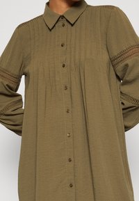 Olivgrünes Hemd mit Knöpfen, das einen kragenartigen Halsabschluss, vertikale Falten und Spitzenakzente an den Ärmeln aufweist. Weicher, leichter Stoff.