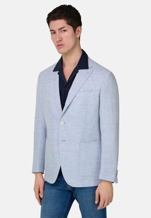 Junger Mann trägt einen hellgrauen texturierten Blazer über einem dunkelblauen Hemd mit Kragen und blauen Jeans, steht vor einem schlichten Hintergrund.