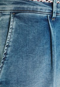 Modré denimové džíny mají světlé praní, jemnou strukturu a viditelné švy. V pase jsou přítomny sklady, které zdůrazňují design.