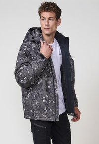 Chaqueta acolchada en tonos de negro y gris con un patrón texturizado, que presenta una capucha, cierre de cremallera y puños ajustables.