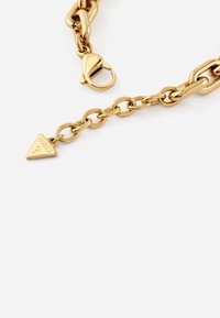 Collana in metallo dorato con catena a maglie di dimensioni alternative. Presenta una chiusura a granchio e un ciondolo triangolare inciso con "GUESS".
