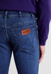 Džinsai su tamsiai mėlynu skalbimu ir lygiu paviršiumi. Galinės kišenės viduje yra rudos odos Wrangler logotipo pleistras ir siuvinėtos detalės.