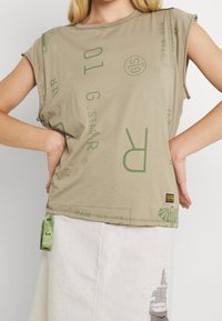 Woman wearing a beige graphic print sleeveless top with rolled cuffs and a light-colored skirt with a distressed patch.