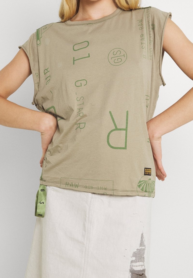 Woman wearing a beige graphic print sleeveless top with rolled cuffs and a light-colored skirt with a distressed patch.