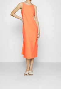 Bezramenné midi šaty v jasně oranžové barvě. Ušité z hladké tkaniny, mají volný střih a boční rozparek. Kombinované se světlými neutrálními sandály.