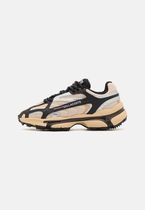 Beige und schwarze Sportschuh mit Mesh- und synthetischen Overlays, klobiger Sohle, schwarzen Schnürsenkeln und Lacoste-Logo auf dem Seitenpanel.