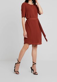 Robe brune à manches courtes avec une ceinture à la taille, pr ésentant une coupe décontractée et un tissu léger et lisse. Assortie avec des talons noirs à lanières.