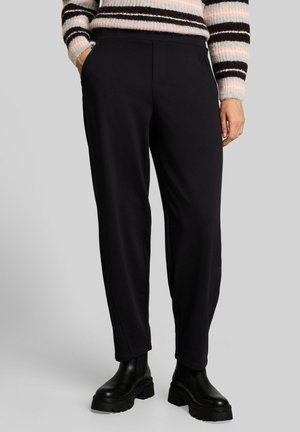 Person, die schwarze gerade geschnittene Hose, schwarze klobige Stiefel und einen rosa-weiß-schwarz gestreiften Pullover trägt, mit Händen teilweise in den Taschen.