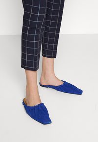 Blå mockaimules med spetsig tå och rynkad elastisk kant, kombinerat med marinblå rutig byxor i cropped-modell med ljusblå ränder.