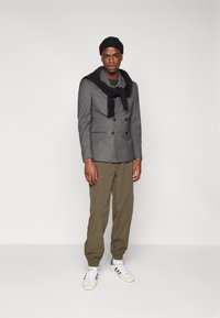 Siv trojni blazer, črna jopica ovita okoli ramen, khaki trenerke in bele superge z črnimi detajli.