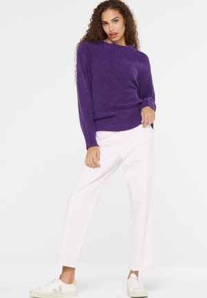 Woman wearing a purple long-sleeve sweater, white wide-leg pants, and white sneakers standing against a plain white background.