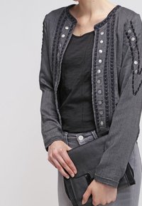 Szara, teksturowana kurtka z dekoracyjnymi guzikami i przeszyciami, noszona na czarnej bluzce, zestawiona z czarną torebką kopertową.