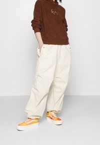 Sudadera con capucha de forro polar marrón con un bolsillo frontal, combinada con pantalones crema de corte holgado. Zapatillas deportivas en tonos naranja brillante y amarillo con suelas blancas.