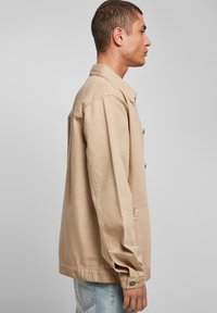 Beige bomullsjakke med avslappet passform, store sidelommer, knappedetaljer og lange ermer, vist fra siden.