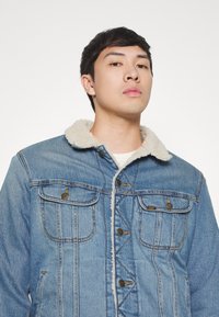 Zilas džinsa jaka ar mīkstu krēmkrāsas mākslīgās aitas kažokādas apkakli, priekšējām kabatām ar flapi un bronzas pogām. Iezīmē klasisku dizainu un šuvju izstrādājumu.